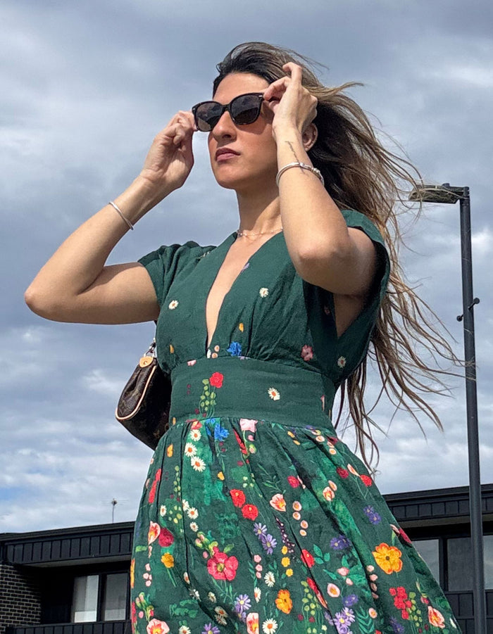 Woman in a green floral devoi dress adjusting sunglasses against a cloudy sky.