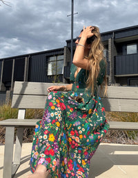 Woman in a colorful floral linen devoi dress sitting on a bench outdoors with a modern building in the background.