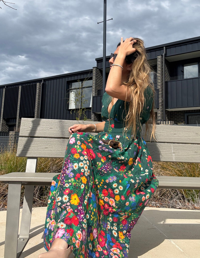 Woman in a colorful floral linen devoi dress sitting on a bench outdoors with a modern building in the background.