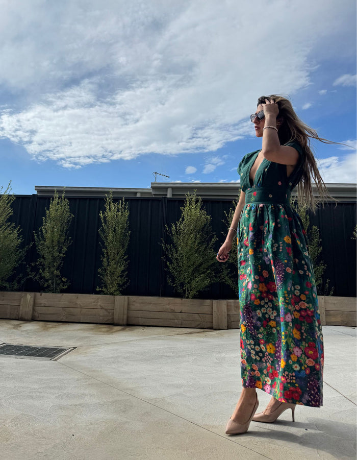 Woman in a colorful floral dress standing outdoors with a blue sky and clouds in the background.