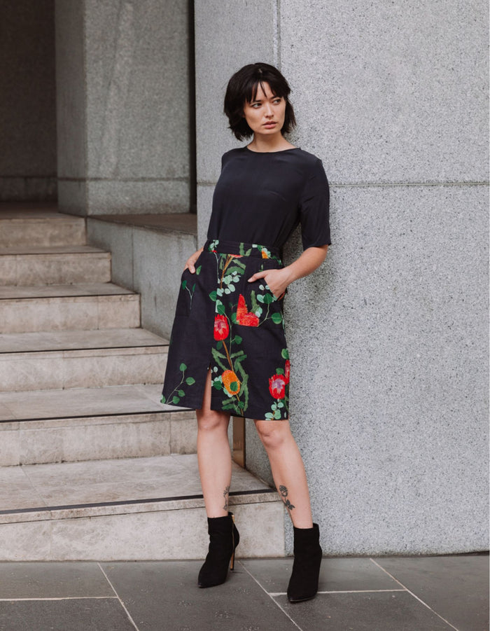 Woman wearing a black DEVOI Maya skirt in Wild Blooms floral print in linen, standing on stone steps.